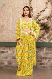 Woman wearing a yellow  printed draped skirt set with a bustier blouse and embroidered cape in Senren's signature  bohemian print against a textured wall.