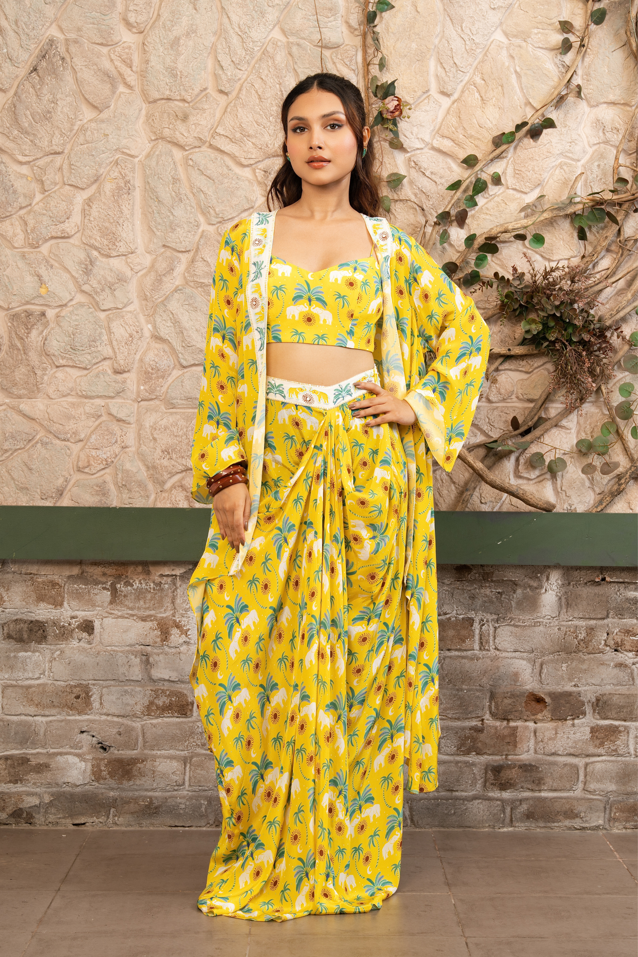 Woman wearing a yellow  printed draped skirt set with a bustier blouse and embroidered cape in Senren's signature  bohemian print against a textured wall.