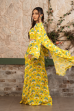 Woman wearing a yellow  printed draped skirt set with a bustier blouse and embroidered cape in Senren's signature  bohemian print against a textured wall.