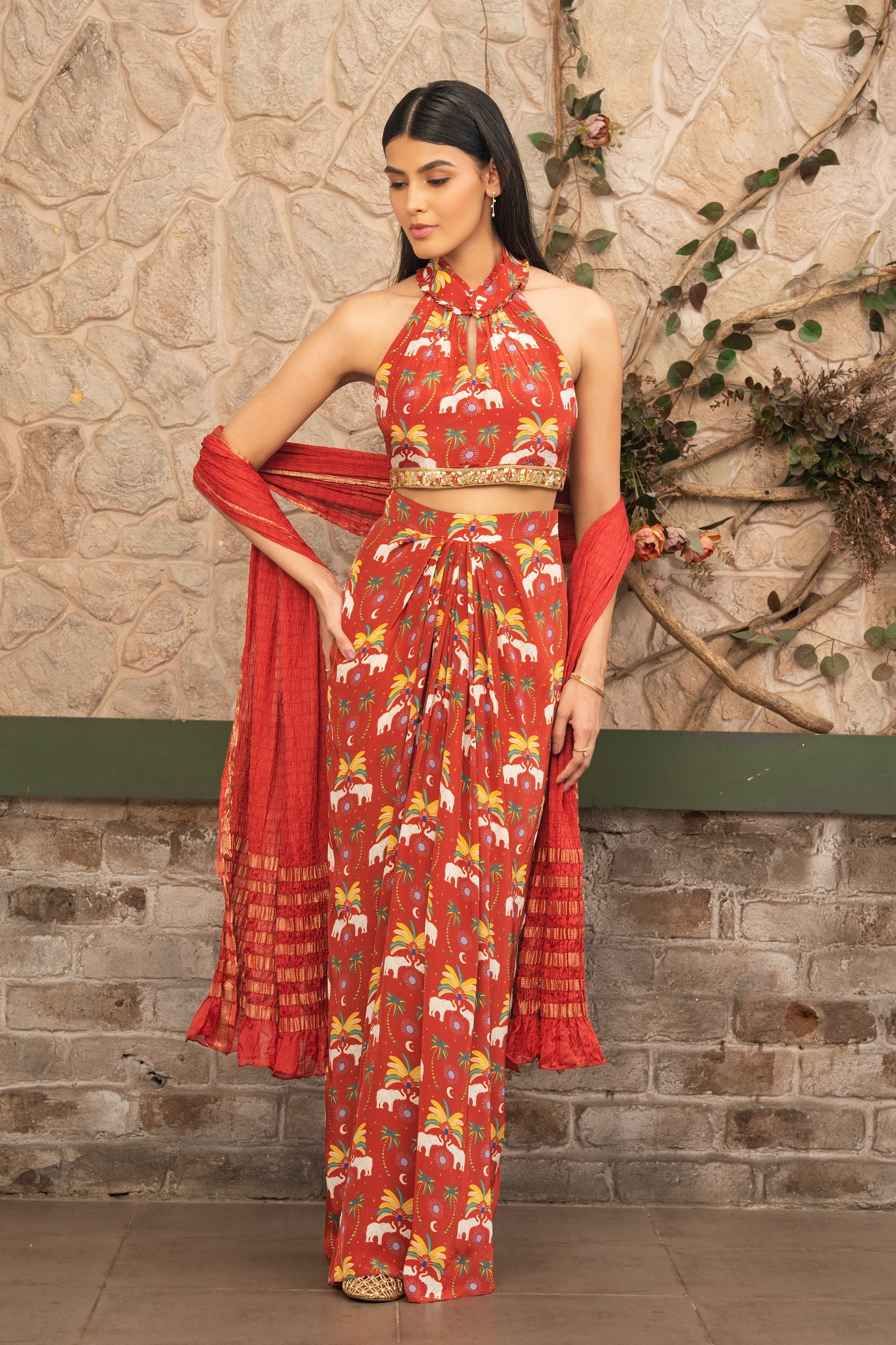 A woman models a red and gold printed halter-neck co-ord set with a matching dupatta, standing against a rustic stone wall with climbing vines.