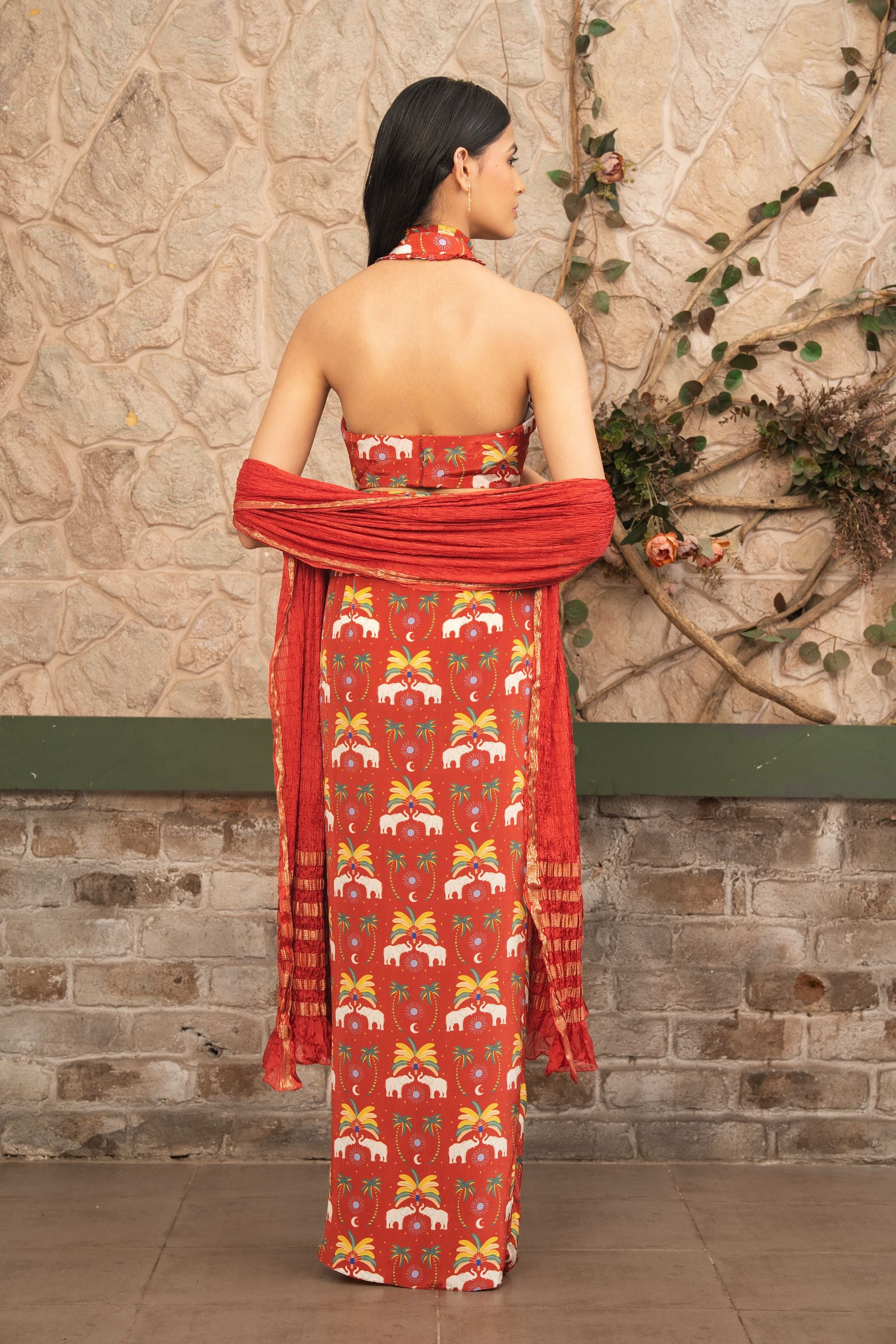 A woman stands with her back to the camera wearing a hand-embroidered  red boho printed halter neck co-ord set with draped skirt and a textured dupatta featuring tropical and elephant motifs by Senren.against a rustic stone wall background.