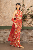 A woman models a red  printed halter-neck two-piece outfit with a matching dupatta, standing against a rustic stone wall backdrop.