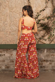 A woman stands with her back to the camera wearing a matching red two-piece set with a white and yellow tropical floral print, consisting of a sleeveless crop top and wide-leg pants, set against a rustic stone wall.