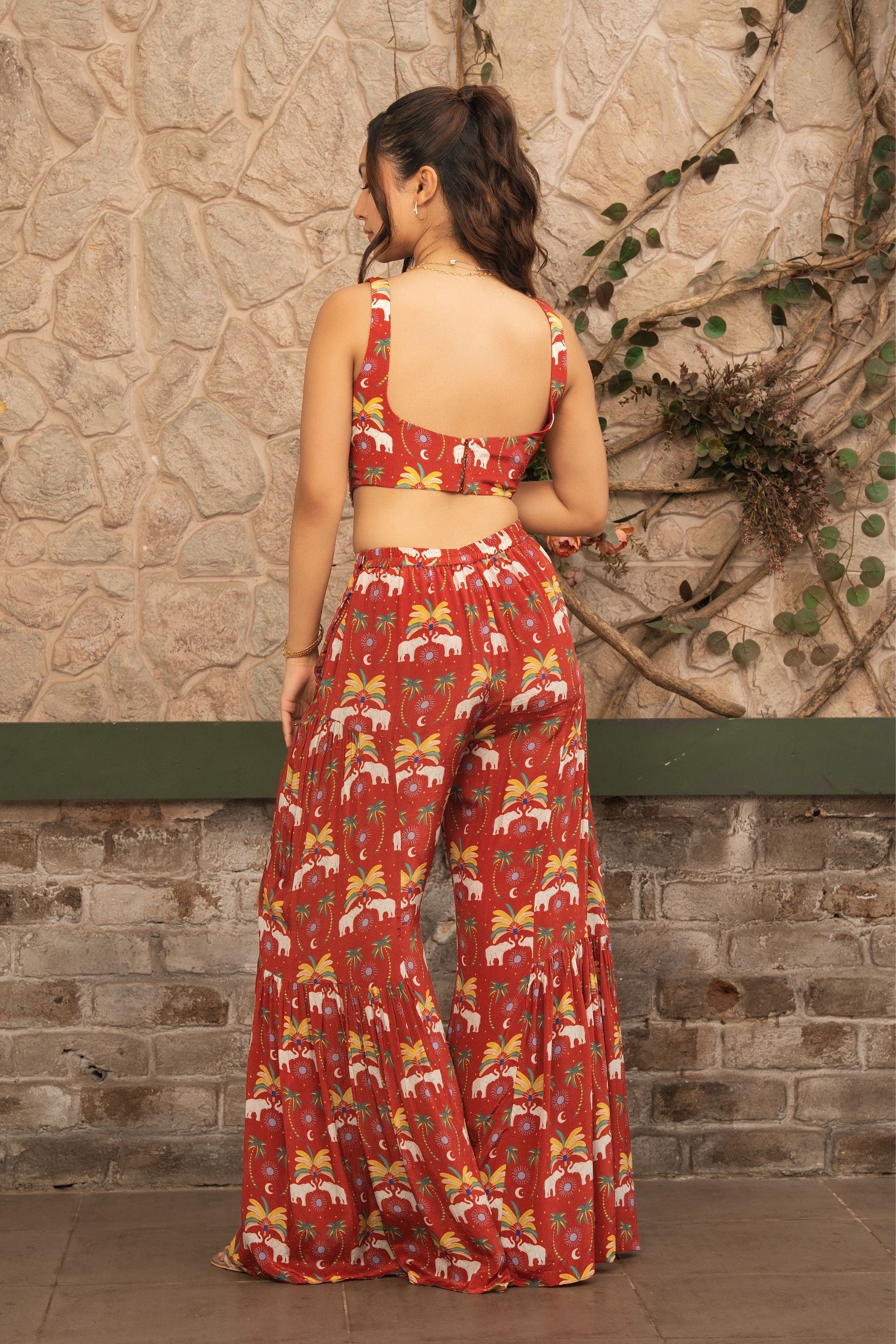 A woman stands with her back to the camera wearing a matching red two-piece set with a white and yellow tropical floral print, consisting of a sleeveless crop top and wide-leg pants, set against a rustic stone wall.