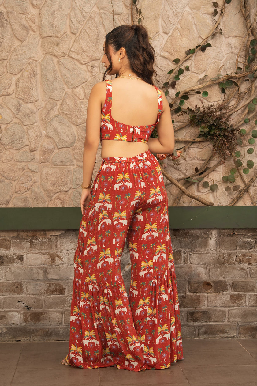A woman stands with her back to the camera wearing a matching red two-piece set with a white and yellow tropical floral print, consisting of a sleeveless crop top and wide-leg pants, set against a rustic stone wall.