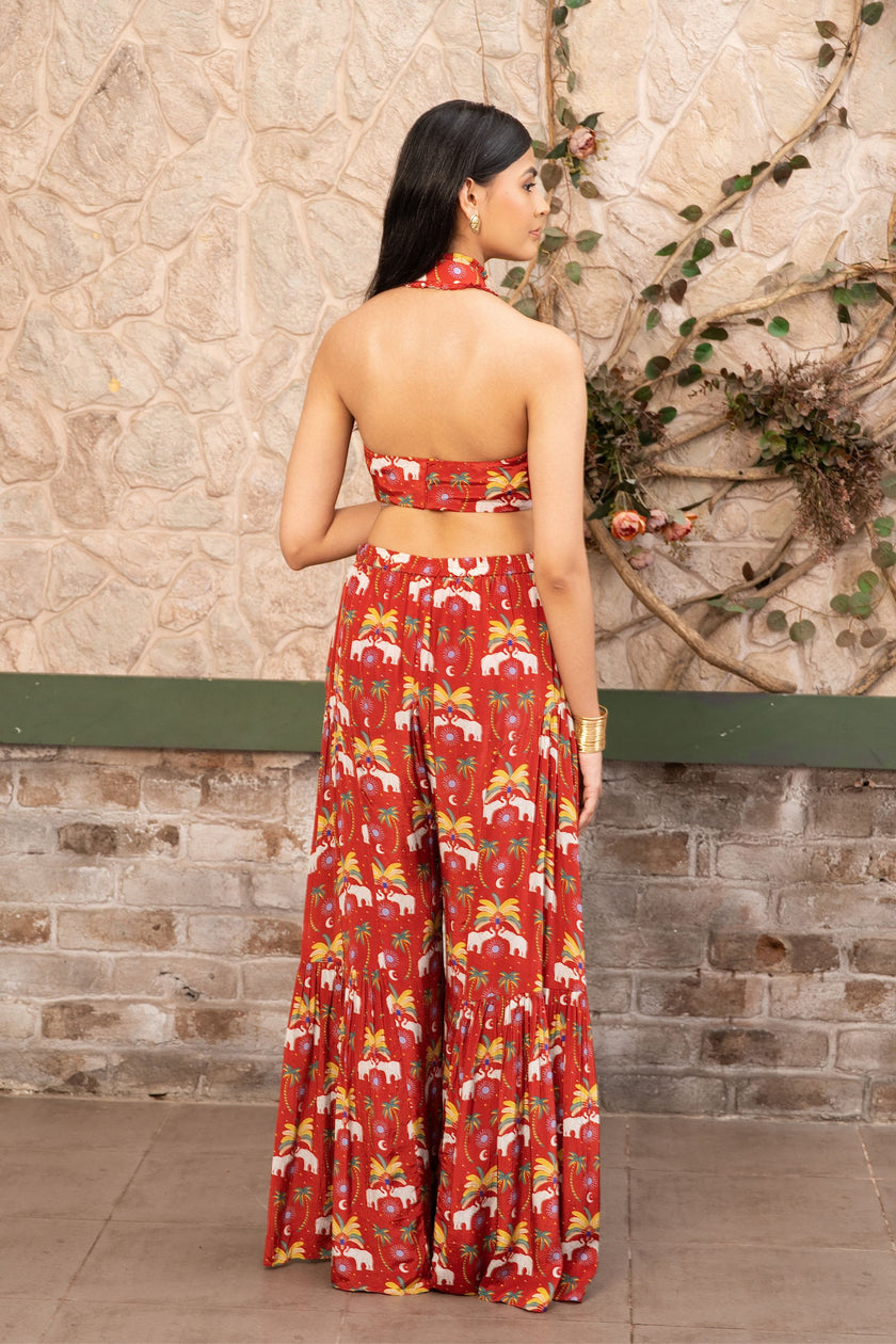A woman stands with her back to the camera wearing a red halter neck printed co-ord set with wide-leg sharara pants in Senren's signature bohemian print.