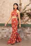 A woman models a red and gold printed two-piece outfit consisting of a halter-neck crop top and a matching flared sharara pants, standing against a textured stone wall with climbing vines.
